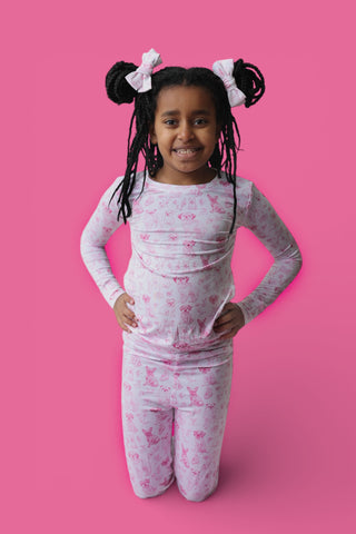 Child wearing long-sleeve white pajama top and pants with pink puppy toile pattern against pink background.
