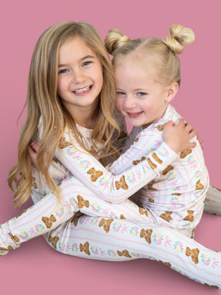 Two young girls wearing white pajamas with bear and floral print against a pink background.
