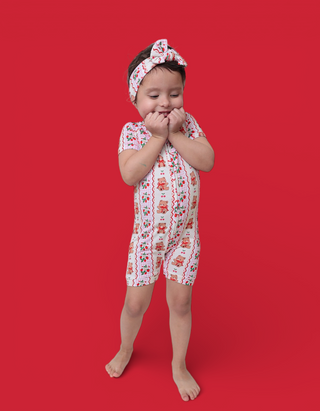 Toddler wearing white short-sleeved romper with red berry and bear print, matching headband, standing against red background.