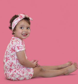 Toddler wearing pink bow-print short-sleeve romper with matching bow headband against pink background.