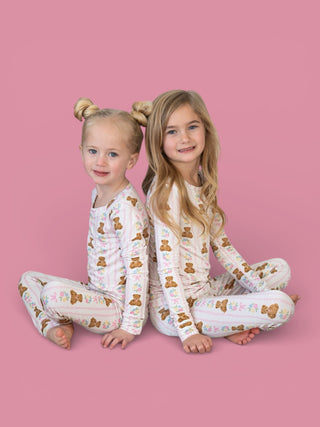 Two children wearing matching cream pajama sets with brown teddy bear and pink floral print, seated against pink background.