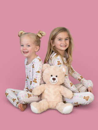 Two girls wearing matching pajamas with bear and floral print, posing with a cream-colored teddy bear against a pink backgrou