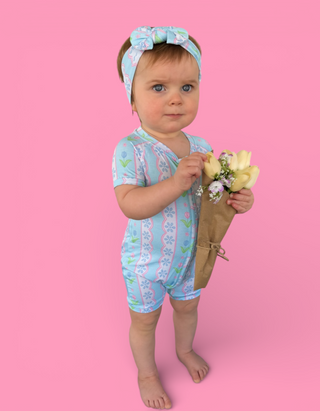 Toddler wearing light blue short-sleeve romper with geometric tulip print and matching headband, holding yellow flowers.