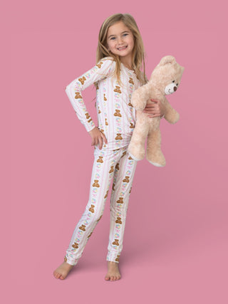 Girl wearing white long-sleeve pajama set with bear and floral pattern, holding matching plush teddy bear.