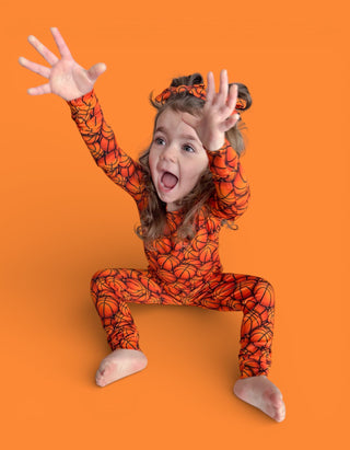 Child wearing orange basketball-patterned pajamas with long sleeves and pants, posing with arms raised against orange backgro