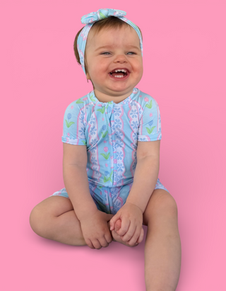 Toddler wearing light blue short-sleeve romper with tulip and geometric print and matching bow headband.