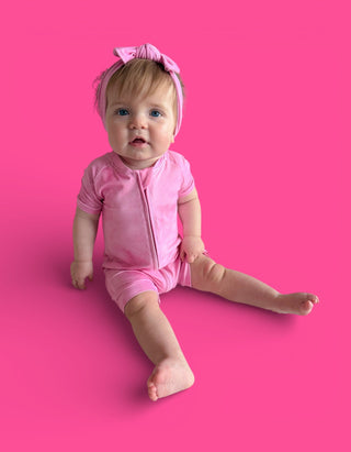 Infant in pink short-sleeved romper with matching bow headband against pink background.