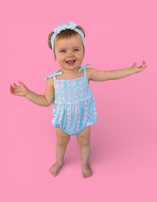 Toddler wearing light blue bubble romper with tulip print, matching headband, and tie shoulder straps against pink background