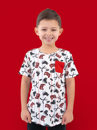 Child wearing white short-sleeve tee with red and black hockey sticks and pucks print, red pocket on chest.