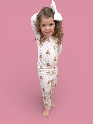Young child wearing white pajamas with brown bears and floral print, matching hair bow, standing against pink background.
