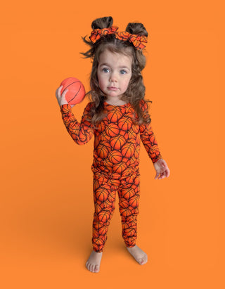 Toddler wearing orange basketball-print pajamas with matching hair bows, holding a small basketball.
