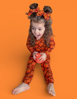 Child wearing orange basketball-print pajama set with matching hair bows against orange background.