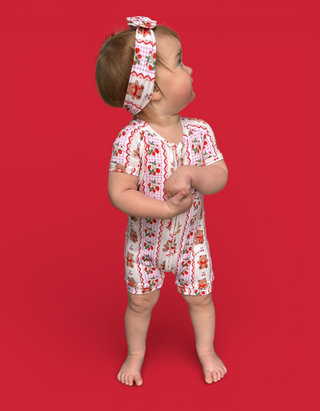 Toddler wearing white romper with red berry and bear print, matching headband, standing against red background.