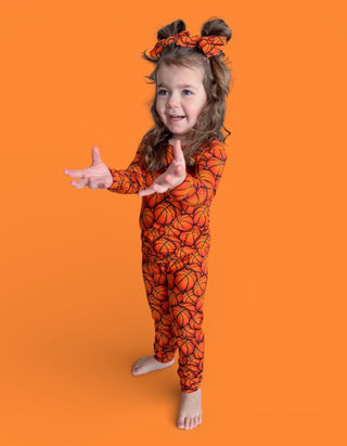 Young child wearing orange basketball-print pajamas with matching hair bows against an orange background.