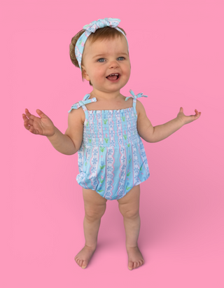 Infant in light blue bubble romper with floral and geometric pattern, matching bow headband and shoulder ties.