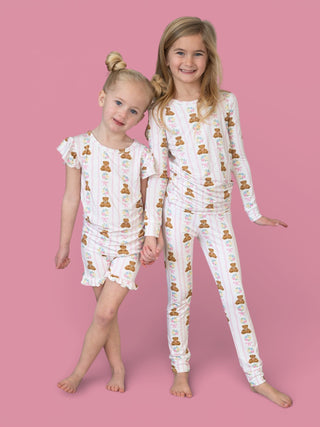 Two young girls wearing white pajamas with brown teddy bears and pink flowers printed throughout, one in short sleeves and sh
