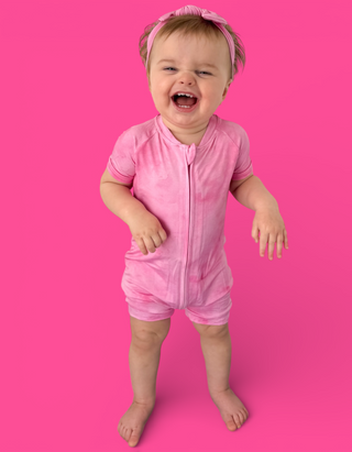 Toddler wearing pink short-sleeved romper with front zipper and matching pink headband on bright pink background.