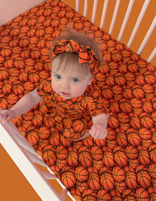 Orange crib sheet with repeated basketball pattern, shown on infant in striped crib with matching headband.