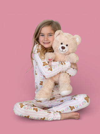 Child wearing cream pajamas with bear and floral print, holding matching plush teddy bear against pink background.