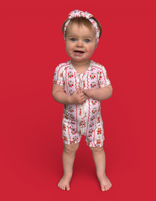 Toddler wearing white short-sleeved romper with red berry and bear print, matching headband, standing against red background.