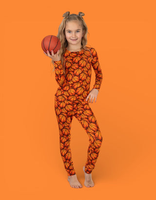 Child wearing orange basketball print pajama set holding a basketball ball against orange background.