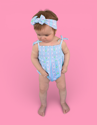 Toddler wearing light blue bubble romper with tulip print, shoulder ties, and matching headband with bow on pink background.