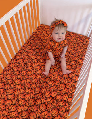 Baby lying on orange crib sheet with basketball pattern and matching headband in white crib with striped bumper.