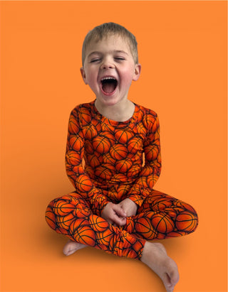 Child wearing orange long-sleeve pajama set with basketball pattern, seated against orange background.