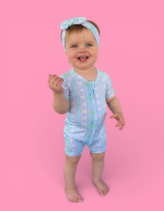Toddler wearing blue short-sleeve romper with tulip and geometric pattern, matching headband, standing against pink backgroun