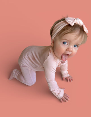 Infant wearing light pink bow headband and matching long-sleeved outfit on coral background.
