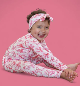 Toddler wearing pink and white bow-printed romper with matching headband against pink background.