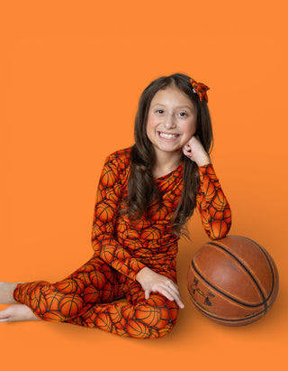 Child wearing orange basketball-print pajamas with matching hair clip, sitting next to a basketball.