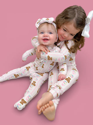 Two toddlers wearing matching white pajamas with brown bears and floral print, with coordinating hair bows against pink backg