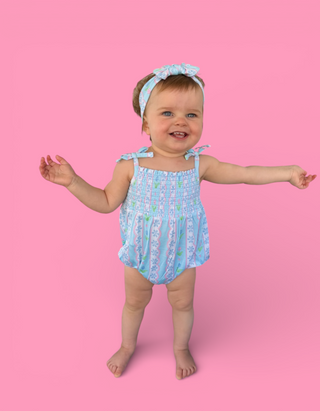 Toddler wearing light blue tulip-print bubble romper with matching headband and shoulder ties against pink background.